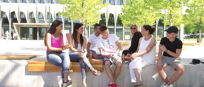 Seven students sitting on a bench chatting and laughing at the plaza infront of the main building of University of applied siences